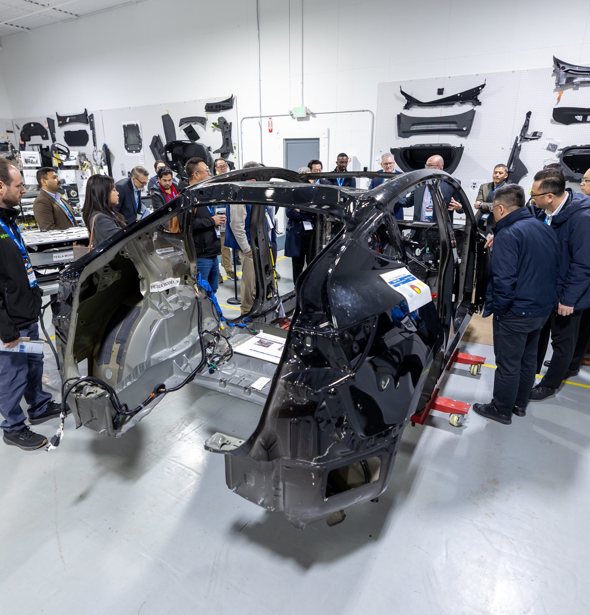 A group of people gather around a partially disassembled black car body in an automotive teardown facility. The stripped vehicle frame is mounted on supports, with exposed wiring and structural components, while additional car parts are displayed on wall-mounted panels in the background.