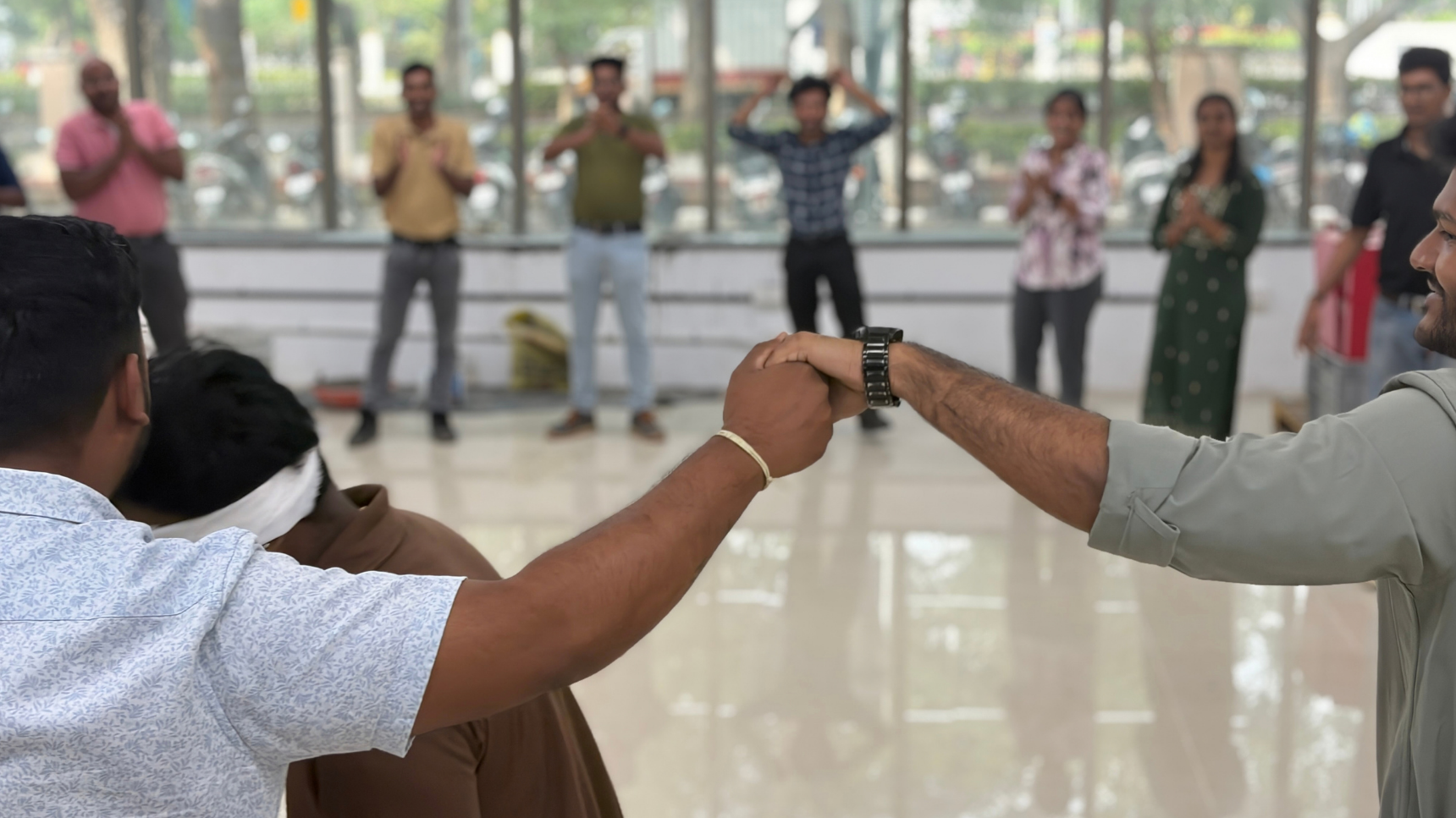 A group of people participates in a team-building activity indoors, with two people in the foreground clasping hands while others stand in a circle in the background.