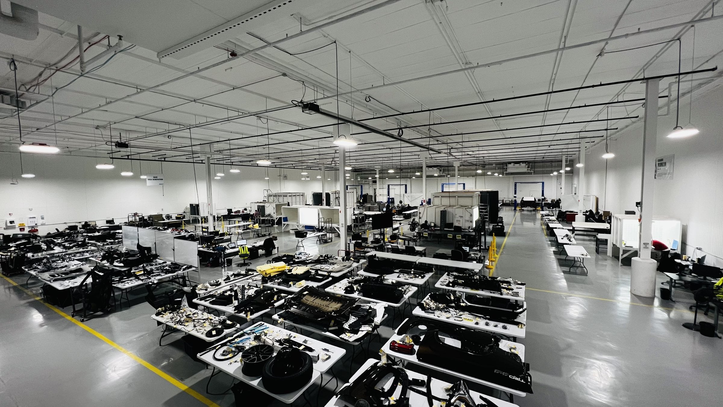 A large, brightly lit automotive teardown facility with rows of tables covered in disassembled vehicle components, workstations, equipment, and computer monitors arranged across the spacious floor.