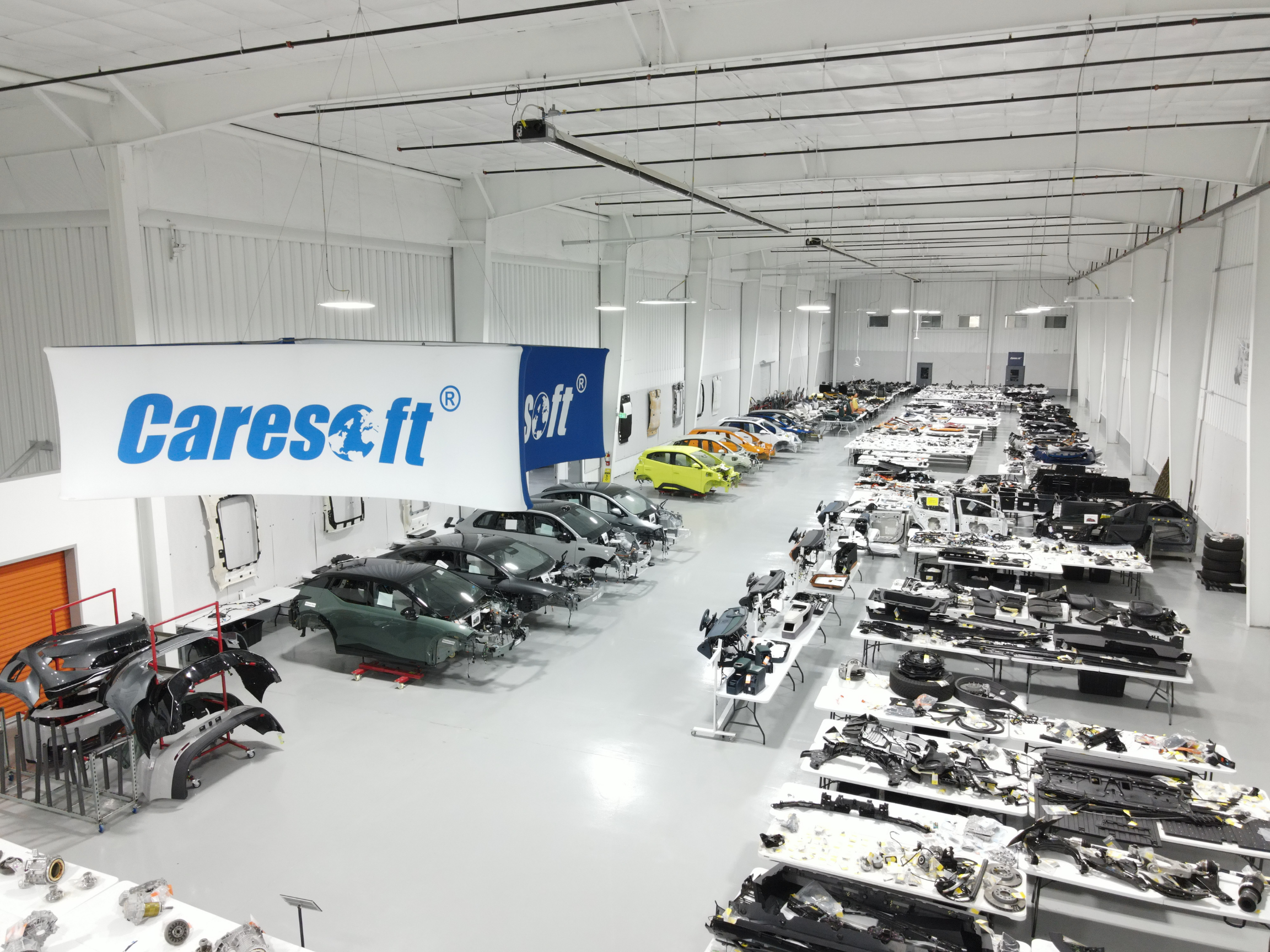 A large, brightly lit automotive parts facility with rows of disassembled car bodies, bumpers, panels, and components arranged on tables and racks. A large “Caresoft” sign hangs prominently in the foreground.