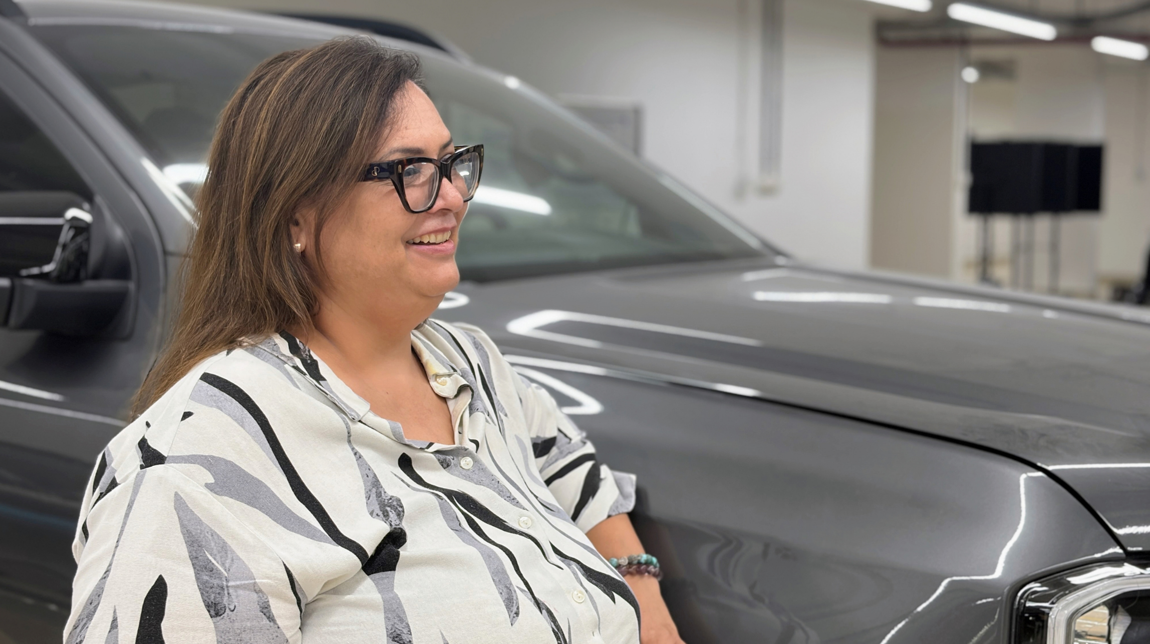 A woman wearing glasses and a patterned blouse smiles beside a dark gray vehicle inside an automotive facility.