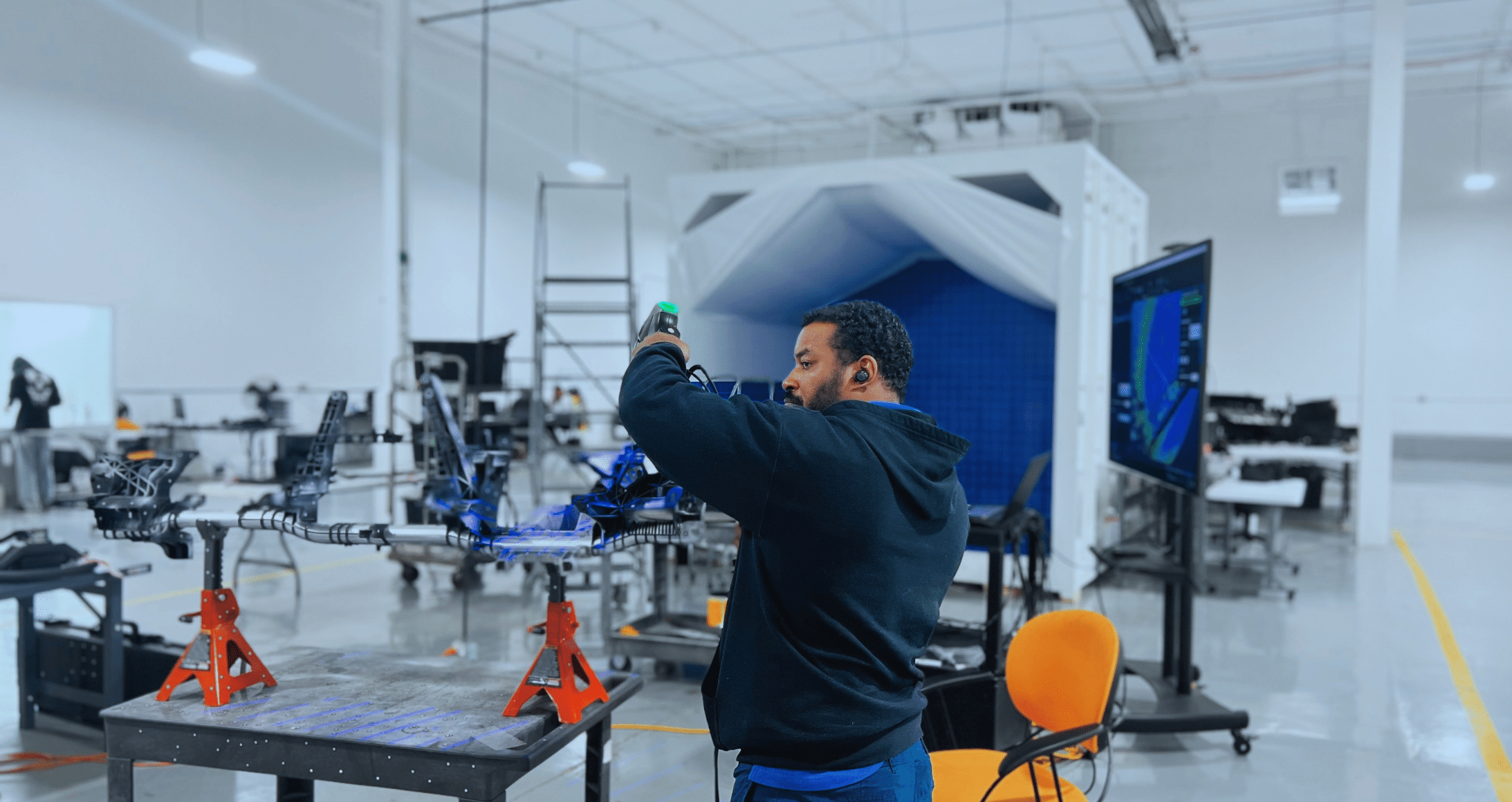 Technician uses a handheld device to inspect equipment in a modern technology laboratory, with workstations, machinery, and a digital display in the background.
