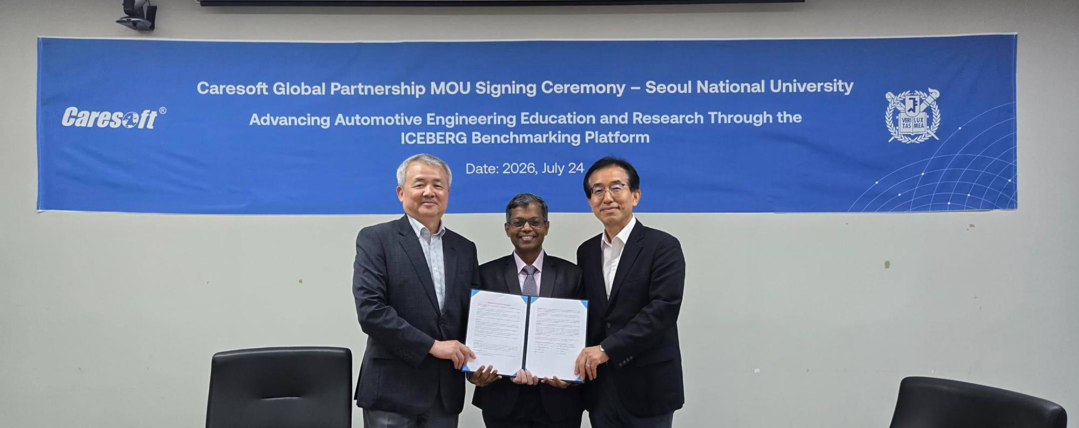 Three men in suits stand together, holding a signed document in front of a blue banner used for a signing ceremony.