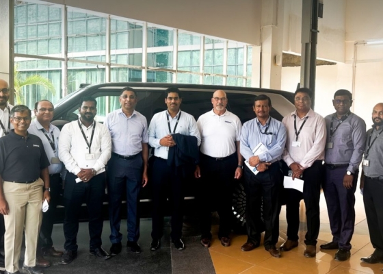 Group of 15 professionals posing in a showroom next to a dark SUV, dressed in business casual attire and name badges.